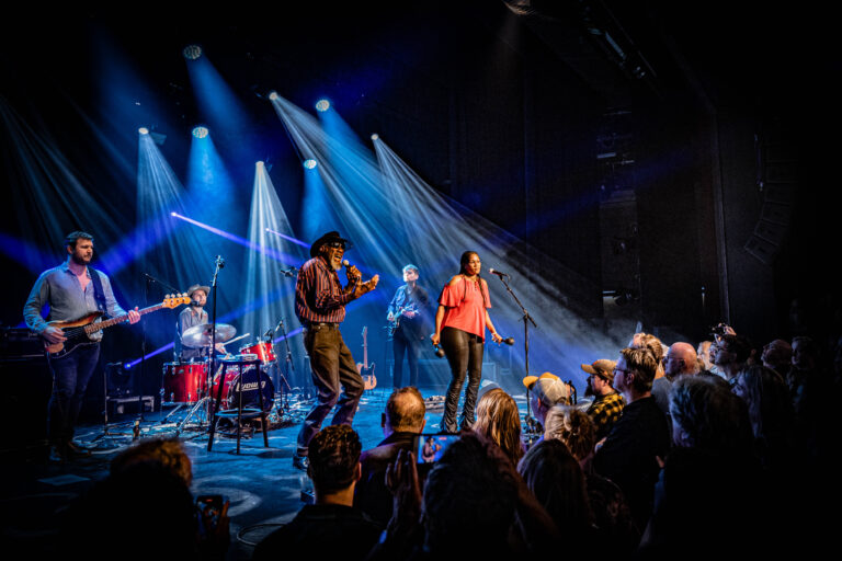 Een band optredend op het podium met een enthousiaste menigte, onderstreept door kleurrijke verlichting en een dynamische sfeer.