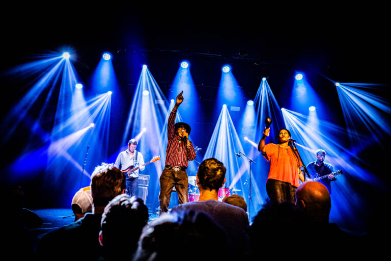 Een muziekgroep performt op het podium met kleurrijke lichteffecten en een enthousiast publiek.