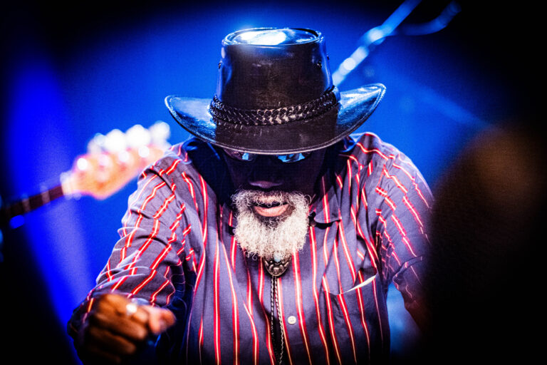 Robert Finley met een hoed en een zonnebril op. Hij draagt een hip gestreepte blouse.
