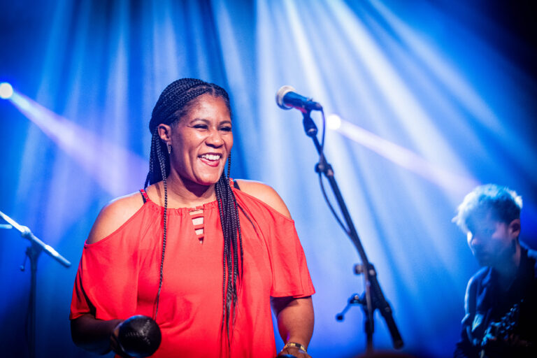 Een glimlachende vrouw in een rode top staat op een podium met verlichting en een microfoon.