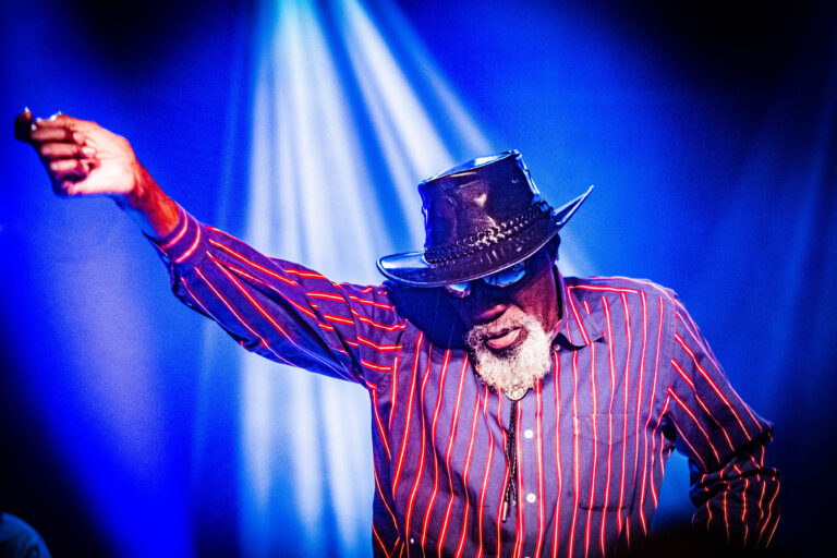 Een man in een gestreepte shirt en hoed staat op een podium met één arm in de lucht, omringd door blauwe lichteffecten.