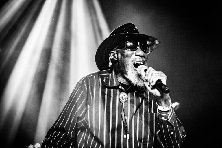 Een oudere man in een zwart-witte outfit zingt gepassioneerd in een behulpzaam verlichte omgeving.