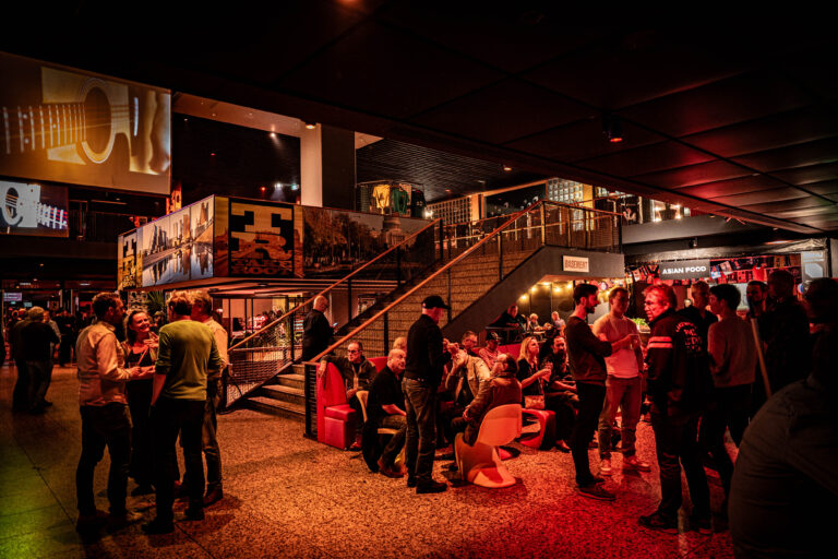 Een druk bezocht café met mensen die aan het socializen zijn, banken en een trap op de achtergrond.