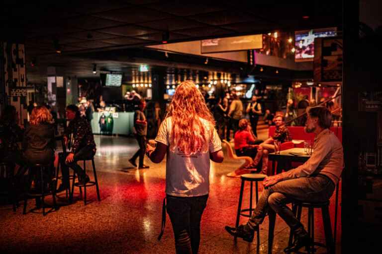 Een persoon met lang, golvend haar loopt door een drukke bar met sfeerverlichting en mensen aan tafels.