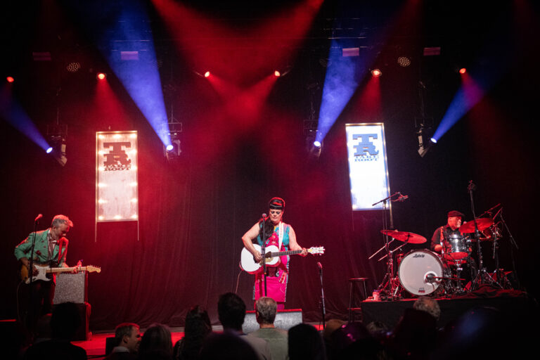 Een muzikant in een kleurrijke outfit speelt gitaar op een podium met schijnwerpers en een drumpartij.