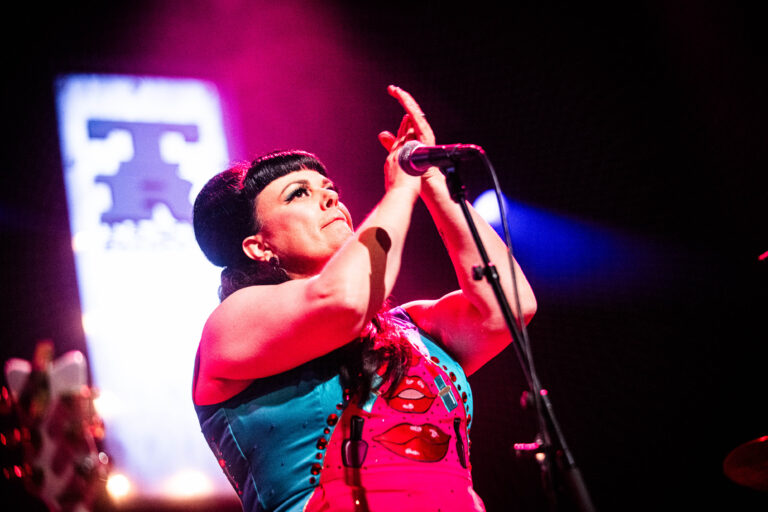 Een zangeres in een kleurrijk outfit staat op een podium, terwijl ze met beide handen een gebaar maakt onder de lichten van een concert.