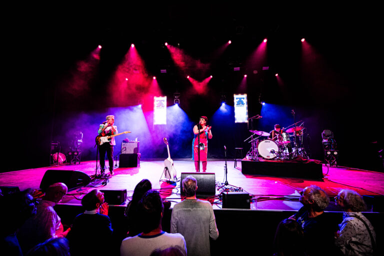 Een live band speelt op een podium met kleurrijke lichten en een enthousiast publiek.