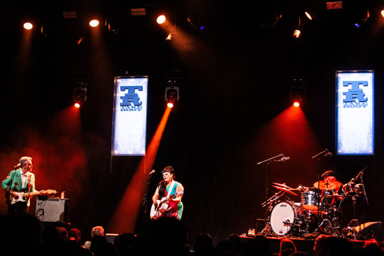 Twee muzikanten spelen live op een podium met kleurrijke verlichting en een banner op de achtergrond.