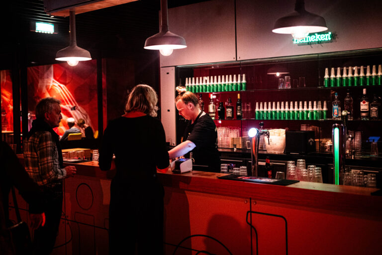 Een bar met een bartender die een drankje bereidt voor een klant, omgeven door warme verlichting en flessen op de achtergrond.