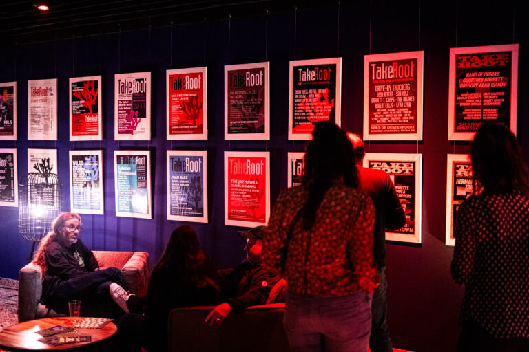 Een ruimte met een paar mensen die naar een muur met ingelijste affiches kijken, in een sfeervolle rode verlichting.