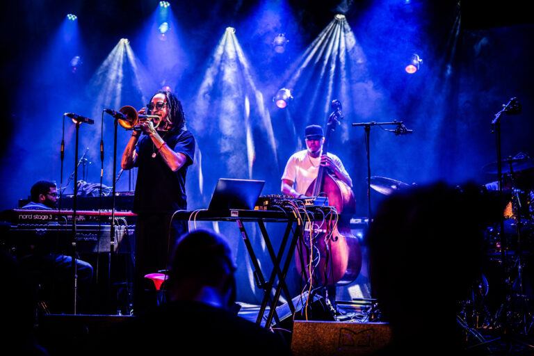 Een muzikant speelt trompet op een podium, omringd door kleurrijke verlichting en andere bandleden.