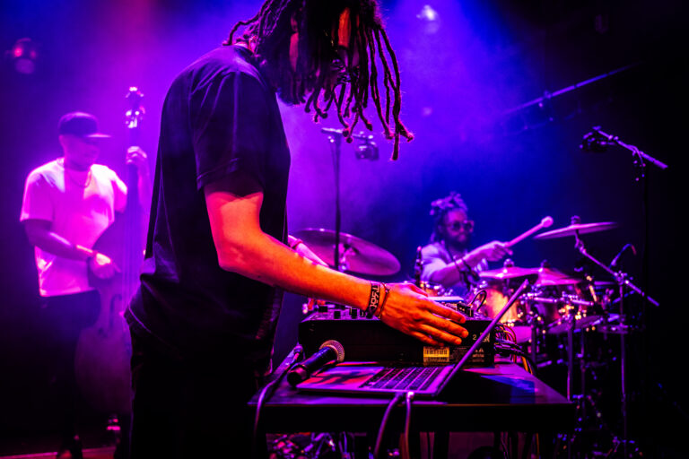 Een muzikant in een zwarte shirt bedient een synthesizer, terwijl een drummer op de achtergrond speelt, met paarse en blauwe verlichting.