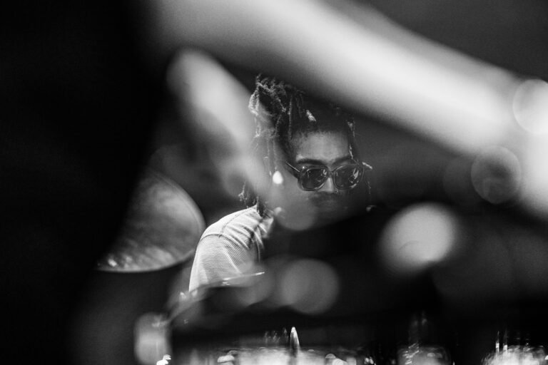 Een muzikant met een zonnebril speelt drums, gefocust op zijn instrument in een zwart-wit afbeelding.