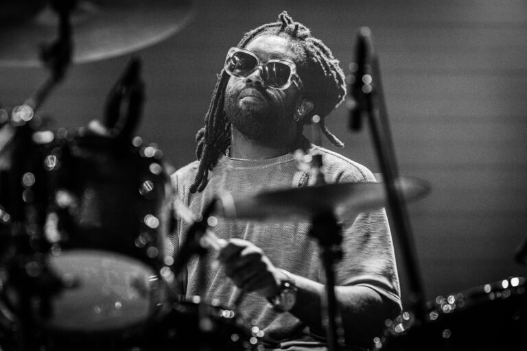 Een drummer met dreadlocks en een zonnebril speelt op een podium.