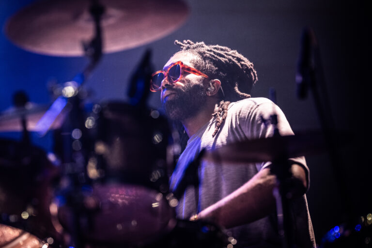 Een drummer in een kleurrijke outfit en rode zonnebril speelt op een podium met belichting.