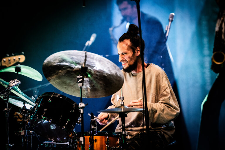 Een drummer speelt enthousiast op zijn drums terwijl een andere muzikant op de achtergrond zichtbaar is.