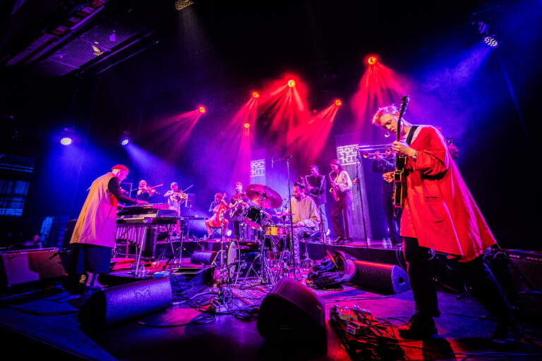 Een band speelt live op een podium met kleurrijke verlichting en diverse instrumenten.