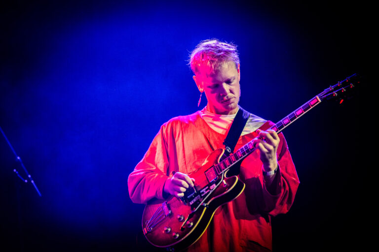 Een muzikant speelt gitaar op een podium met een blauwe en paarse achtergrondverlichting.