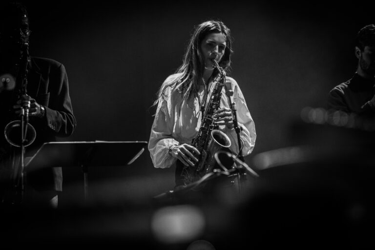 Een muzikant speelt saxofoon op het podium, verlicht door zachte lichten in een zwart-wit setting.