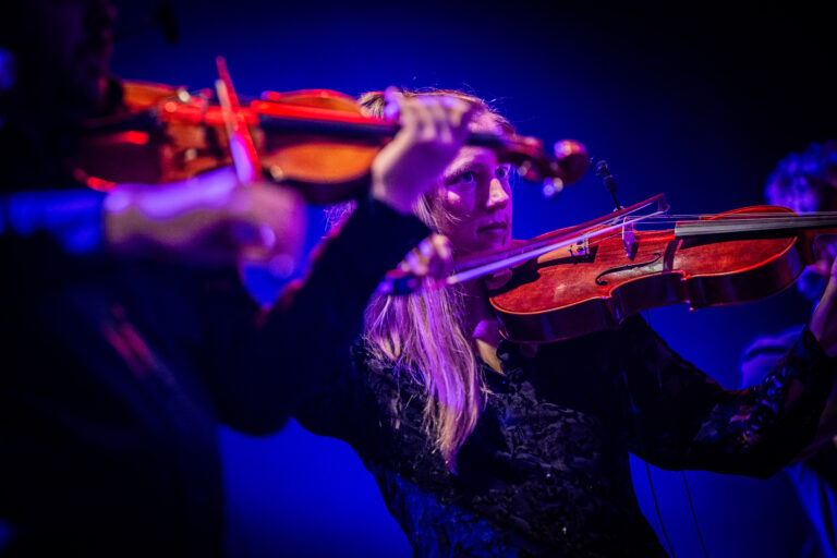 Een close-up van een muzikante die viool speelt op een podium, omgeven door blauwe en paarse verlichting.