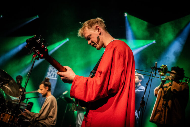 Een muzikant in een rode outfit speelt gitaar op een podium met groene lichteffecten.