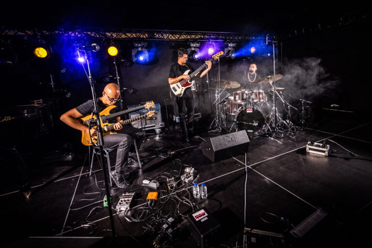 Een trio muzikanten speelt op een podium met kleurrijke verlichting en rookeffecten.