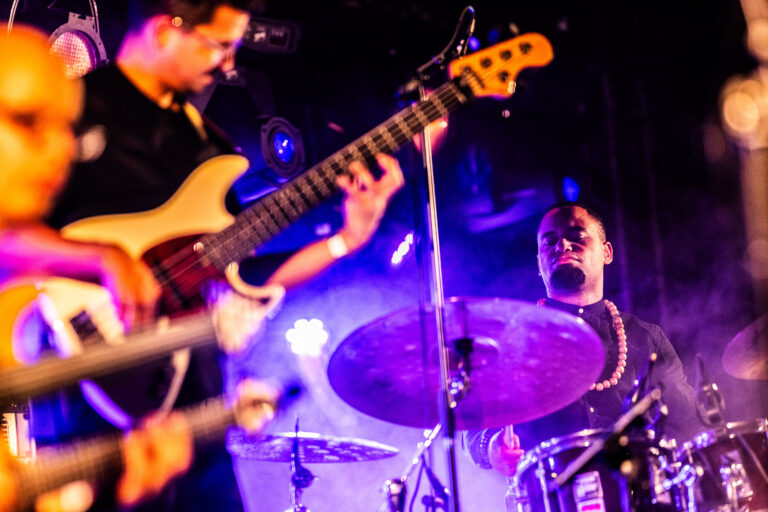 Een muzikant speelt op een elektrische gitaar terwijl een drummer op de achtergrond speelt, omgeven door een paarse gloed van podiumverlichting.