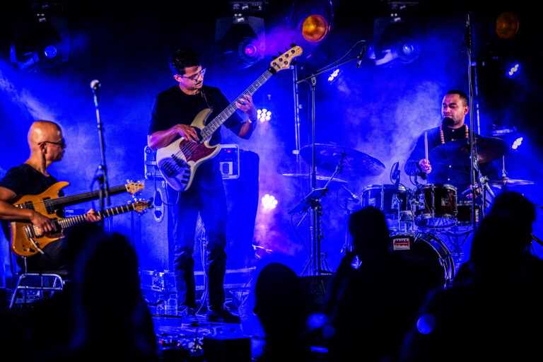 Drie muzikanten spelen op een podium met een kleurrijke lichteffect en een zichtbaar enthousiast publiek.
