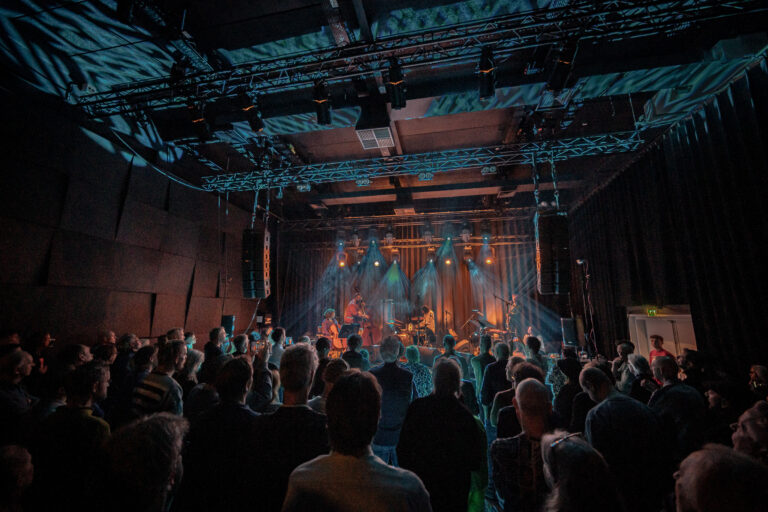 Een druk bezocht concert met een band die op het podium speelt, omgeven door sfeervolle verlichting.