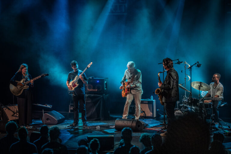 Een live band speelt op een podium met blauwe verlichting, terwijl het publiek aandachtig kijkt.