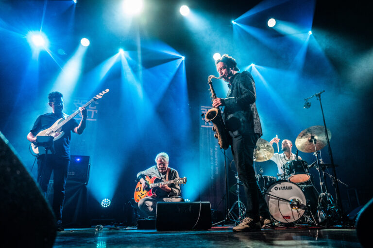 Een band performt live op een podium onder felblauwe lichtstralen.