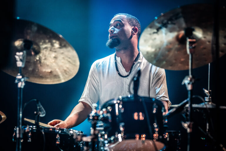 Een drummer in een wit shirt speelt op zijn drums, terwijl blauw licht op hem valt.