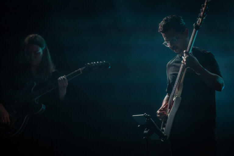 Twee muzikanten spelen gitaar op een podium in een sfeervolle, verduisterde omgeving.