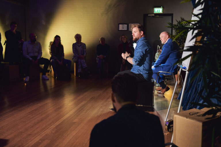 Twee in blauw geklede mannen die spreken voor een groepje mensen.