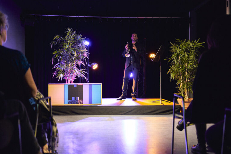 Man met een blaasinstrument op een klein podium met planten eromheen.