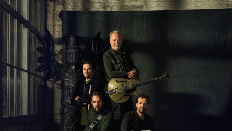 Een band poseert in een donkere setting, met een muzikant die een gitaar vasthoudt.