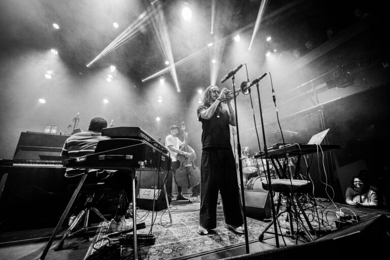 Een muzikant staat op het podium met een microfoon, omringd door andere bandleden en podiumverlichting.