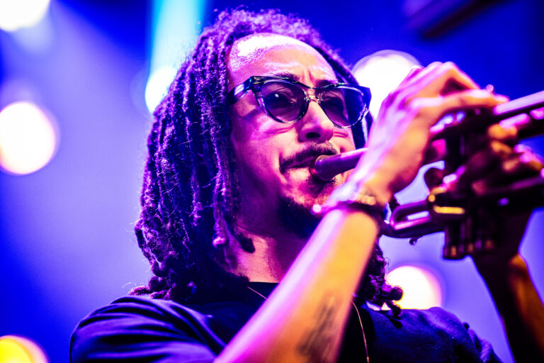 Een muzikant met een trompet, gekleed in een zwart shirt en met een paarse gloed van het podiumlicht.