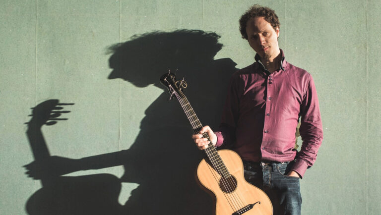 Een man met een gitaar staat voor een muur, terwijl zijn schaduw prominent zichtbaar is.