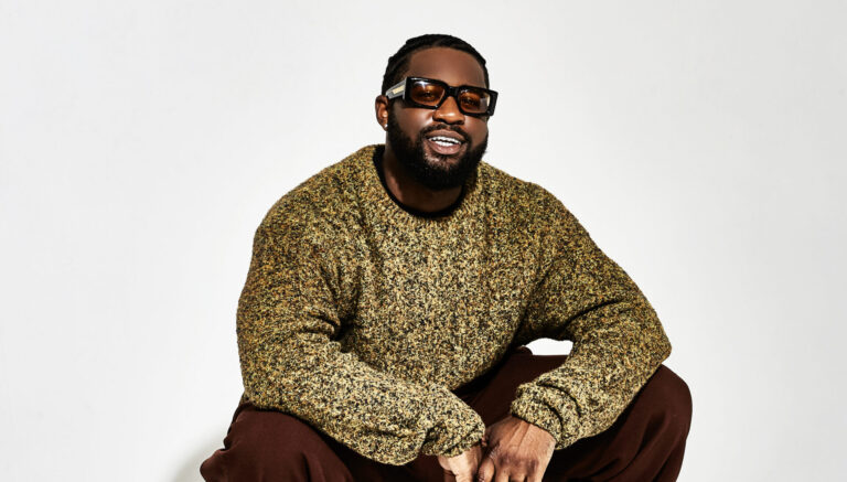 Een man zit in een foto studio met witte achtergrond gehurkt. Hij draagt oldschool hip-hop kleding en ziet er cool uit, inclusief zonnebril en gevouwen handen.