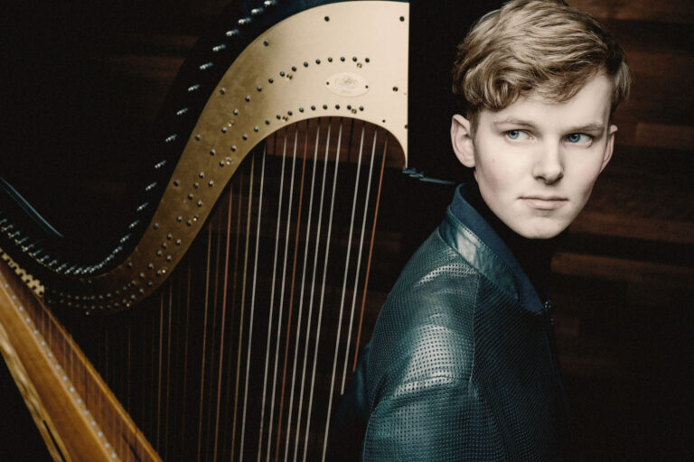 Foto van jongeman die tegen een harp aan leunt en met zijn ogen opzij kijkt.