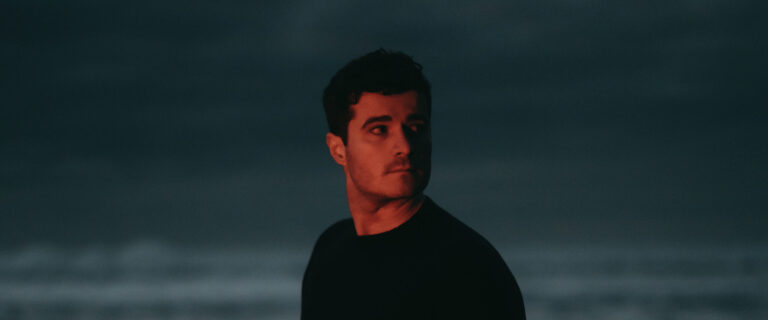 Een man staat op een strand, hij heeft donkere kleding aan en kijkt naar rechts.