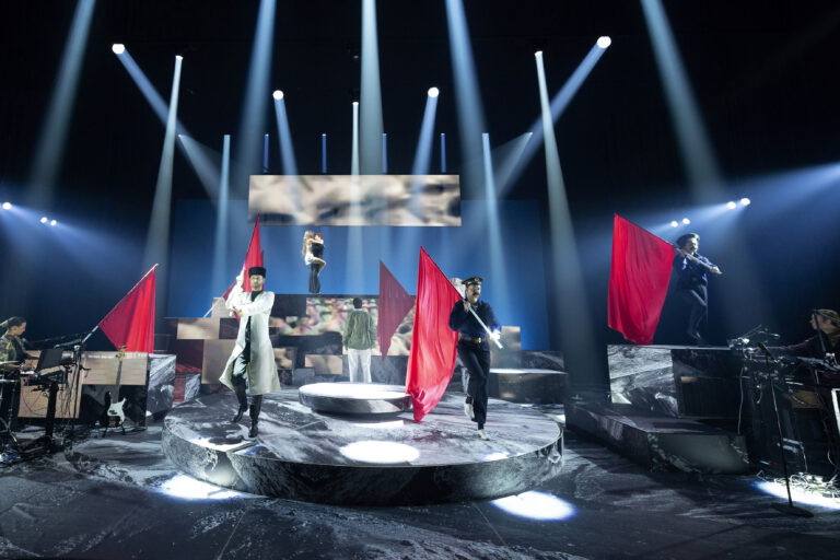 Een podium met performers die rode vlaggen vasthouden, omringd door dramatische verlichting.