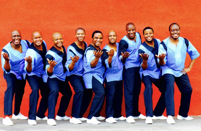 Tegen een oranje muur staan 9 Afrikaanse zangers in flitsende zwart-blauwe pakken