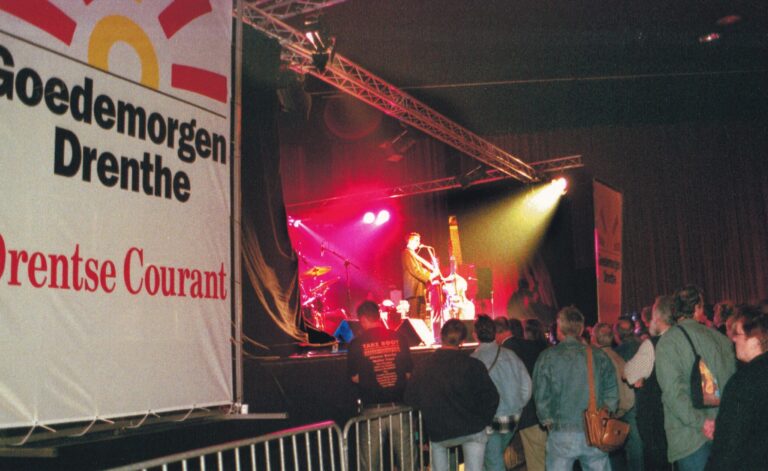 Een band performt live op een podium met kleurrijke verlichting, terwijl het publiek toekijkt.