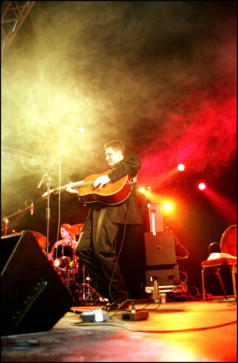 Een muzikant speelt gitaar op een sfeervol podium met kleurrijke verlichting en rookeffecten.