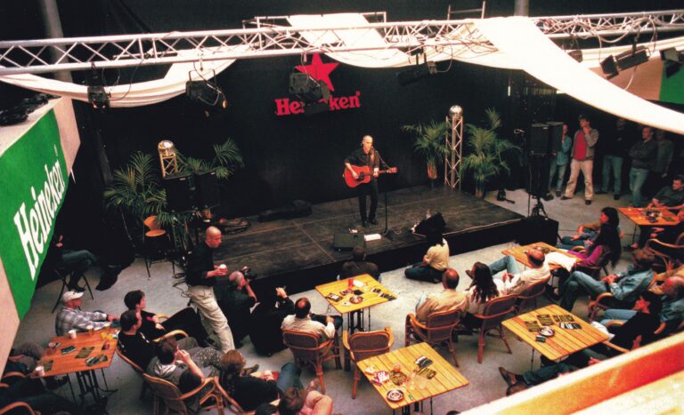 Een muzikant speelt gitaar op een podium, terwijl een publiek in een café-achtige setting toekijkt.