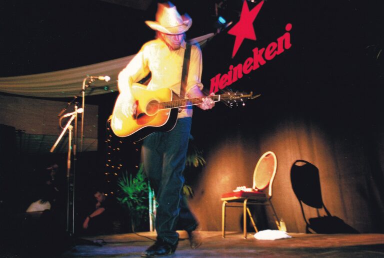 Man met een cowboy hoed die gitaarspeelt met op de achtergrond rode heineken letters.