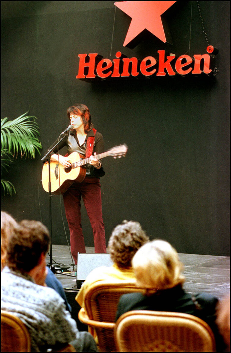 Een zangeres met een gitaar treedt op voor een publiek, met een Heineken-logo op de achtergrond.