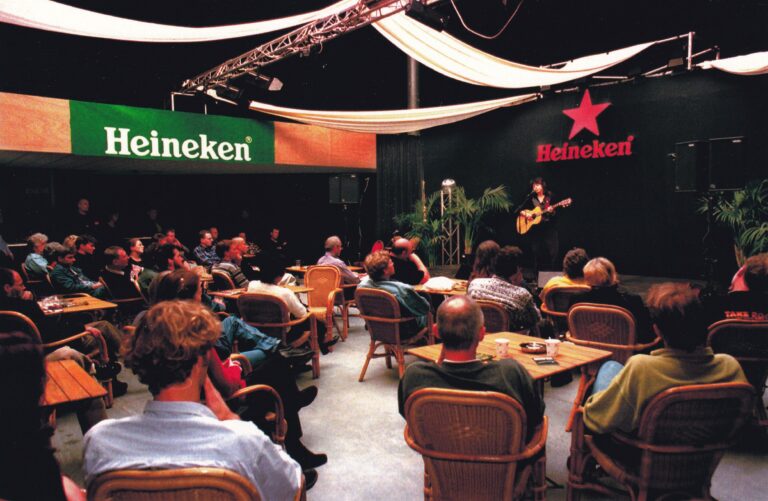 Een klein publiek geniet van een liveoptreden in een sfeervolle zaal met merken als Heineken en Heineken Star op de achtergrond.